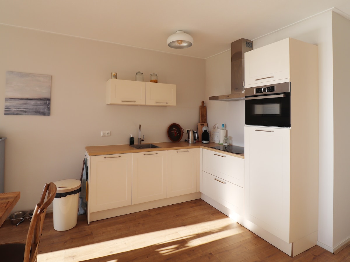 A modern kitchen is equipped with light-colored cabinetry and a wooden countertop. An oven, stovetop, and various kitchen appliances are visible, alongside a dining table and chairs. Natural light enters through a nearby window, highlighting the spaciousness of the area.