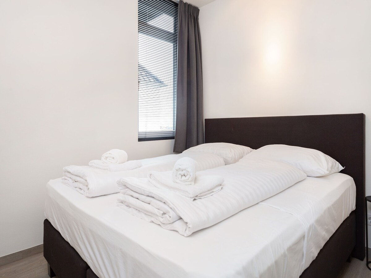 The bedroom features a spacious double bed outfitted with fresh linens and neatly arranged towels. Natural light filters through the window, enhanced by sheer window coverings, while a minimalist décor adds to the modern feel of the room.