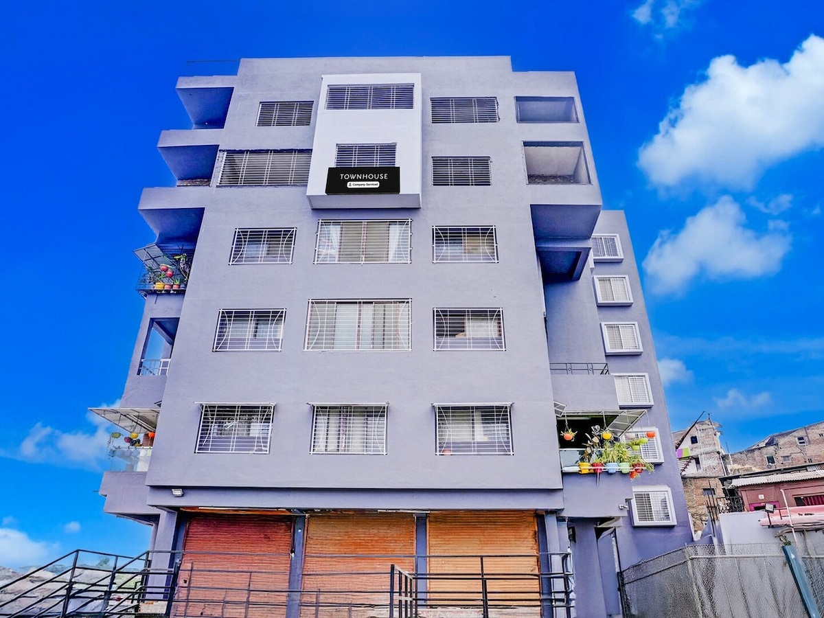 The exterior view of the Townhouse showcases a modern multi-story building. Large windows are visible on each level, providing ample natural light. The front entrance features a gated area, and colorful planters are placed along the balcony railings, adding a touch of greenery.