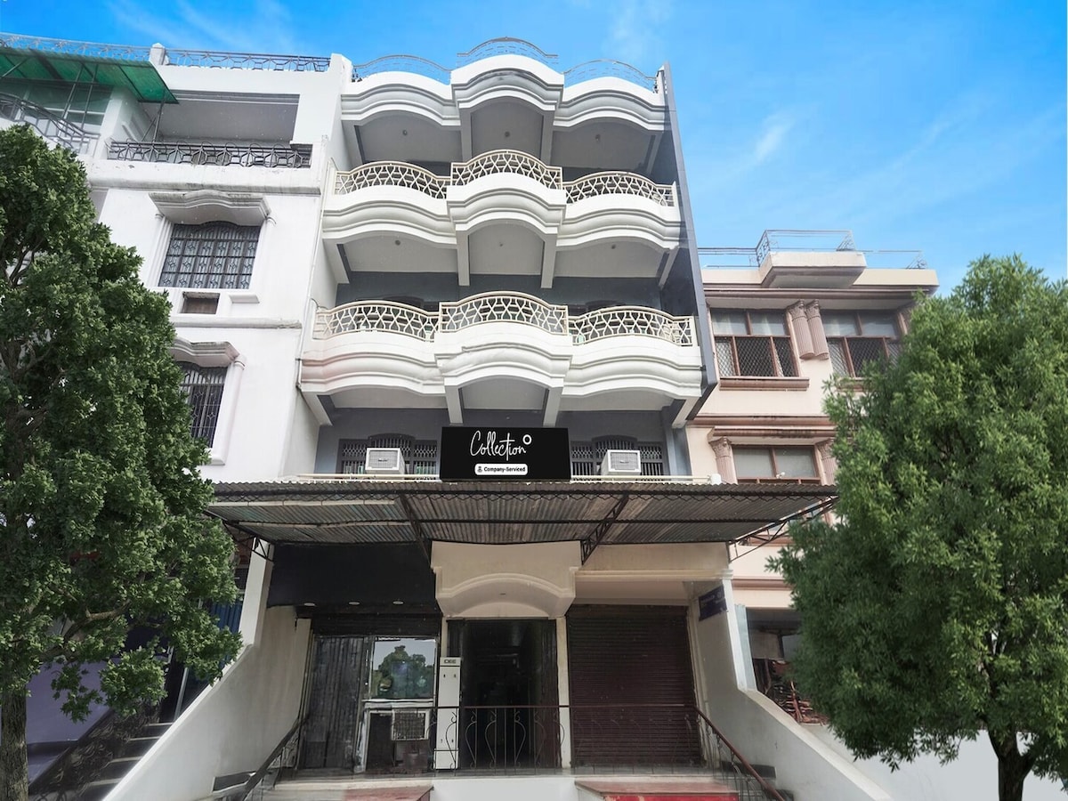 The exterior of the Super Collection O Rajapur Circuit House is shown, featuring a modern structure with multiple balconies and decorative railings. Large windows are visible on each floor, while trees provide greenery on either side of the entrance, which leads to the main lobby.