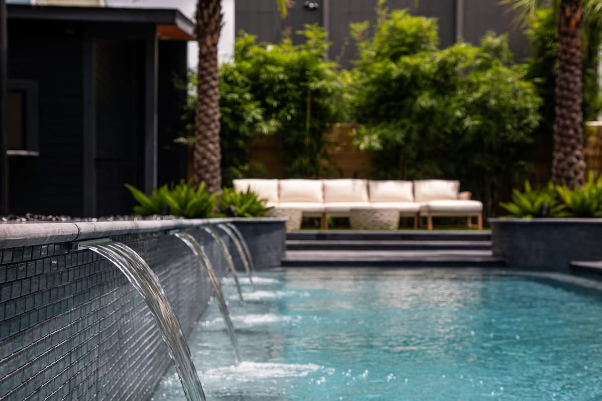 A heated outdoor pool is featured, with gentle water flowing from the tiled edge into the vibrant blue water. Lush greenery surrounds the space, and a comfortable seating area is visible in the background, providing a tranquil spot for relaxation.