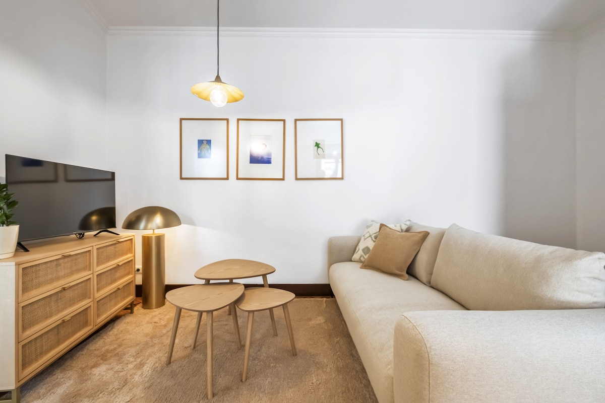 A bright living area features a light-colored sofa and a set of three nested tables. A smart TV is situated on a wooden media console, accompanied by a lamp with a rounded shade. Framed artwork adorns the wall, enhancing the inviting space.