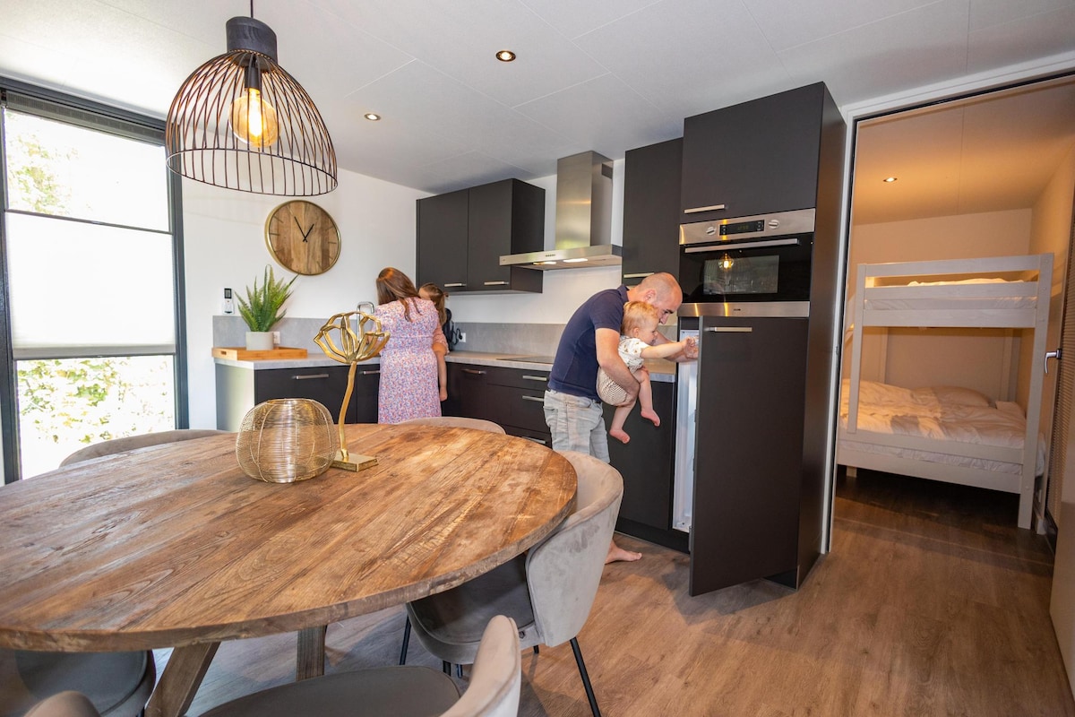 An open-plan kitchen features modern appliances and a spacious dining table, surrounded by upholstered chairs. A family is engaged in preparing a meal, with a child being held. A glass door provides natural light and a view of the outdoors, while a cozy sleeping area is visible through an adjoining door.