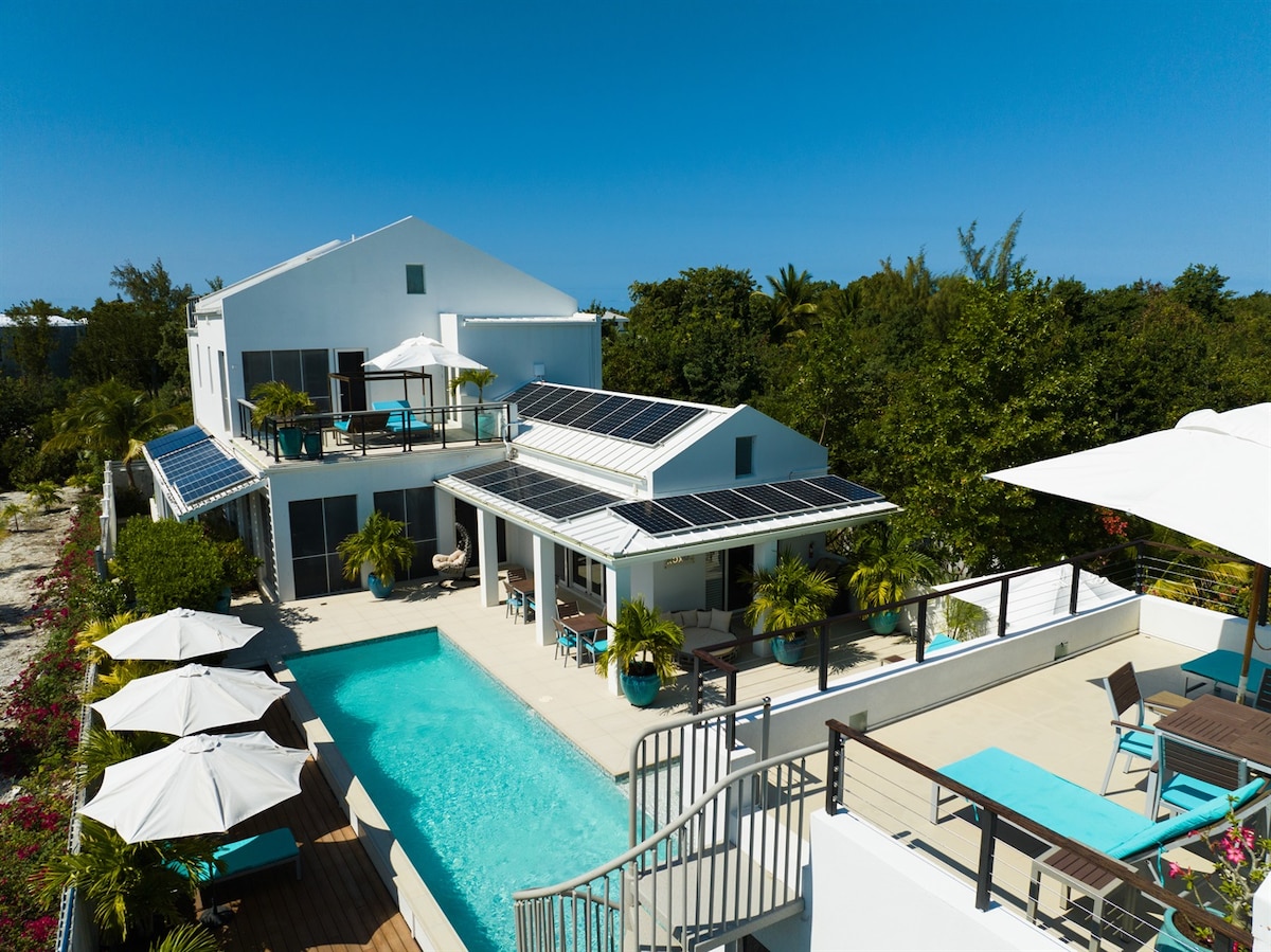 An aerial view of a modern villa features a private pool surrounded by lounging chairs and umbrellas. The structure showcases solar panels on the roof, with vibrant greenery and outdoor seating areas in the background. Clear blue skies enhance the tropical setting.