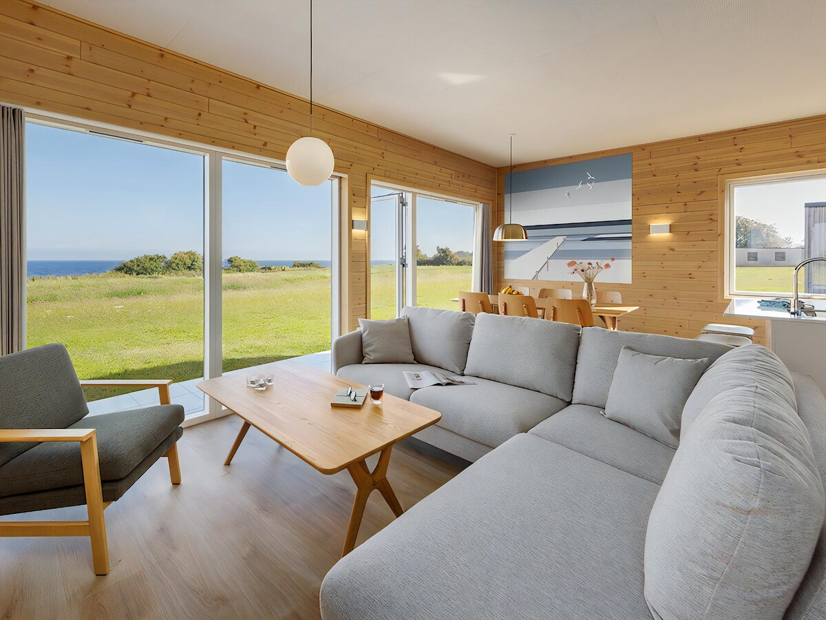 A spacious living area features large windows that provide views of the open landscape and sea. Comfortable seating is arranged around a wooden coffee table, creating a welcoming space for relaxation. Soft natural light fills the room, complemented by wooden accents throughout.
