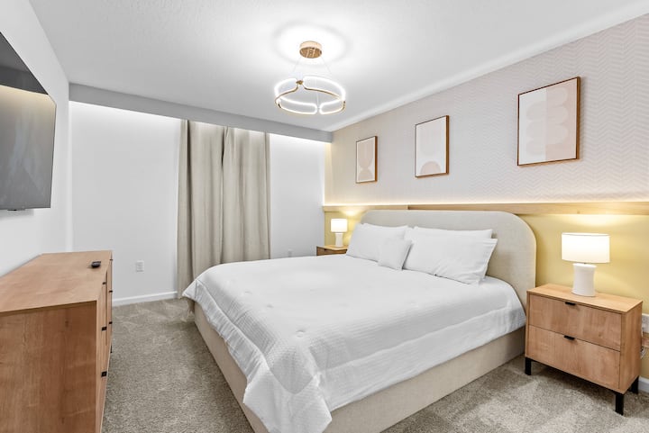 Experience a beautifully styled bedroom with a plush queen bed, crisp linens, warm wood nightstands, and soft lighting. The neutral tones and modern artwork provide a calming atmosphere, ideal for your rest and relaxation.