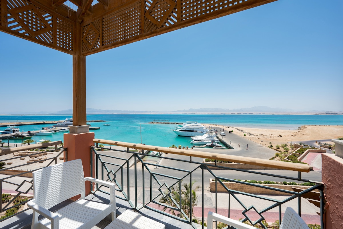 A private terrace features white patio chairs overlooking the marina. The crystal clear waters reflect sunlight, while yachts are anchored in the foreground. The view includes sandy shores in the distance, set against a backdrop of mountains under a clear blue sky.