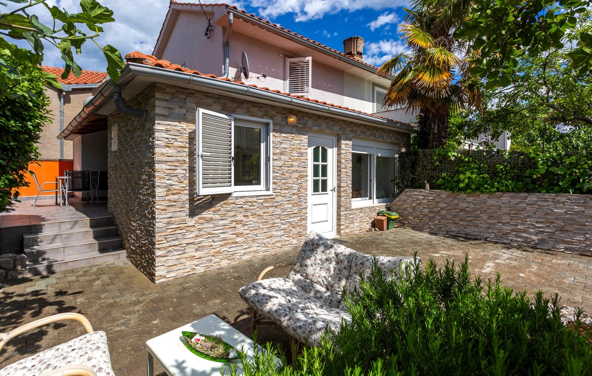 A charming exterior features a stone facade complemented by large windows, allowing natural light to illuminate the space. Outdoor seating is provided on a patio surrounded by greenery, creating a vibrant atmosphere. The property is designed for comfortable outdoor relaxation.