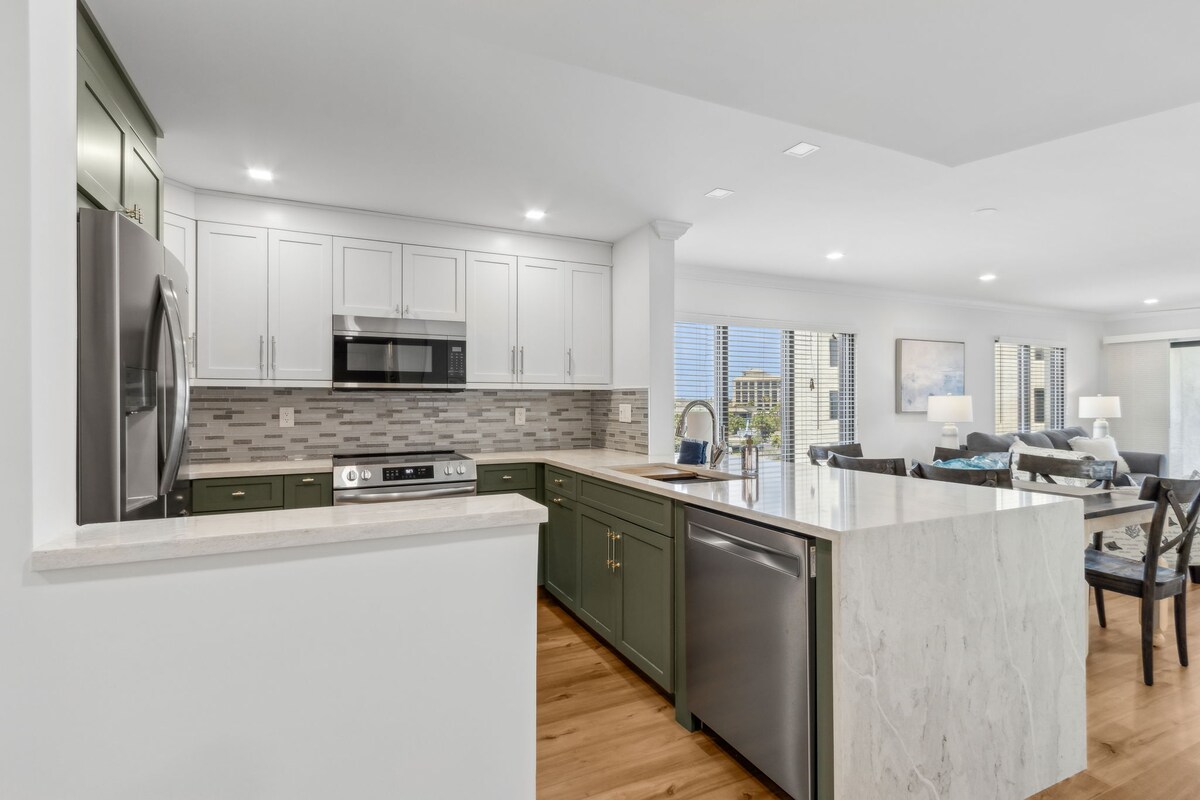 A renovated kitchen features modern cabinetry in dark green with stainless steel appliances, including a refrigerator, microwave, and oven. A spacious island with a light granite countertop provides ample preparation space, while soft lighting illuminates the entire area, enhancing its open design.