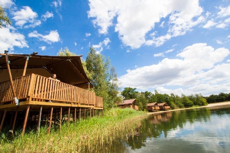 The image captures a waterlodge elevated on wooden stilts, featuring a spacious deck with wooden railings. Lush green grass surrounds the lodge, while the calm water reflects the blue sky dotted with clouds. Additional waterlodges can be seen along the shoreline.