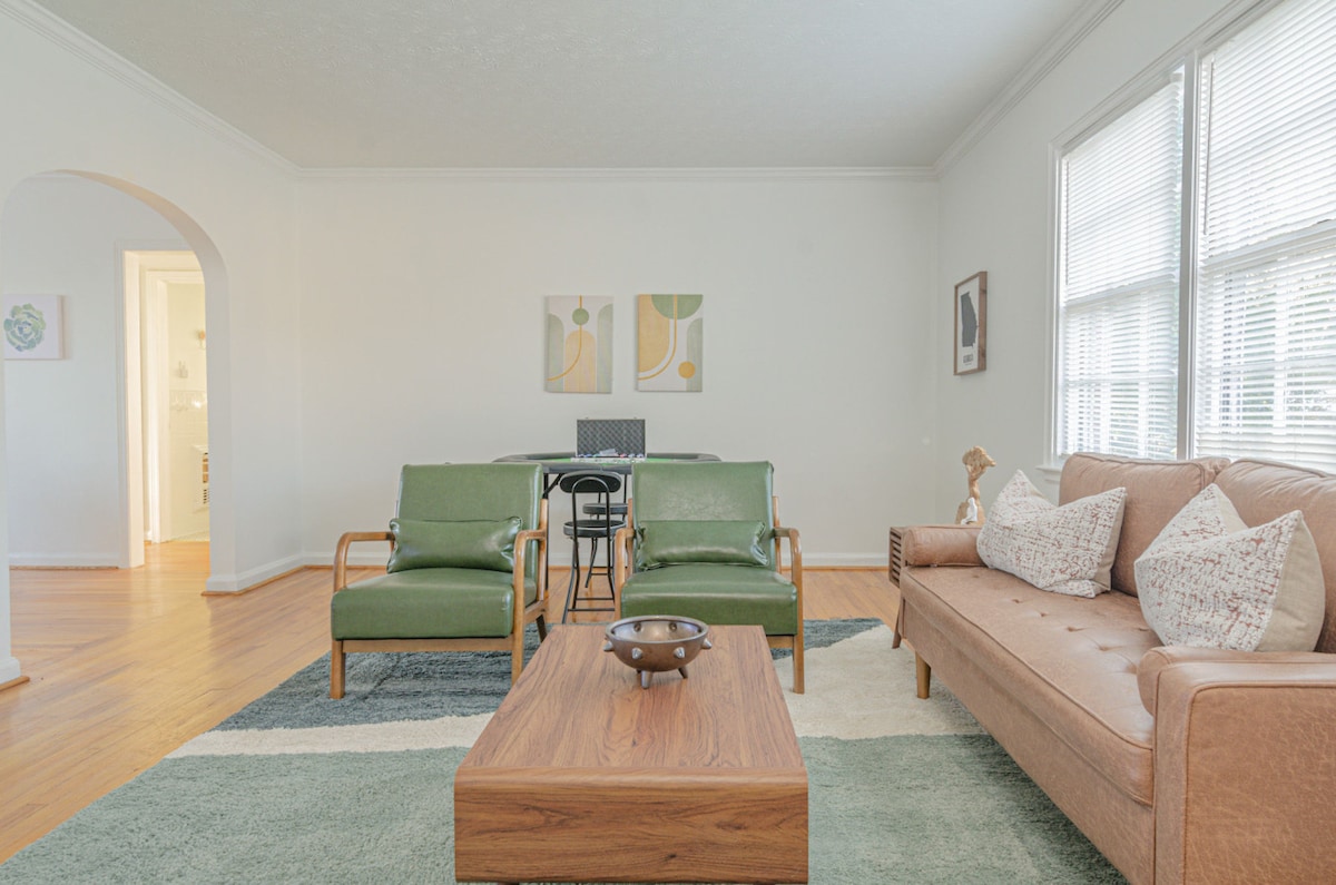 A bright living room features two green armchairs and a tan sofa, offering a comfortable seating area. A wooden coffee table occupies the center, with decorative items placed on it. A dining table is visible in the background, complemented by natural light from the windows.