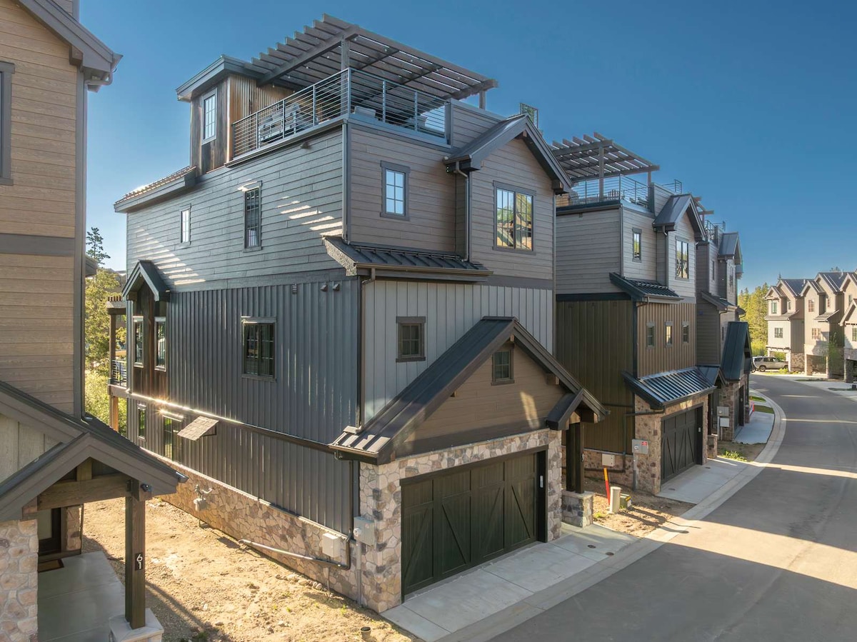 The townhome is positioned on a gentle curve of the road, showcasing its modern exterior featuring a mix of stone and dark siding. The rooftop area is visible, hinting at a spacious outdoor space. Multiple windows reflect the sunlight, enhancing the overall architectural detail.