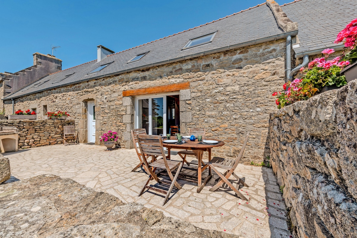 An outdoor terrace features a wooden dining table set with plates and utensils, surrounded by chairs. Colorful flowering plants enhance the stone walls, and large glass doors provide access to the interior. Sunlight brightens the spacious stone-paved area, creating a welcoming environment.