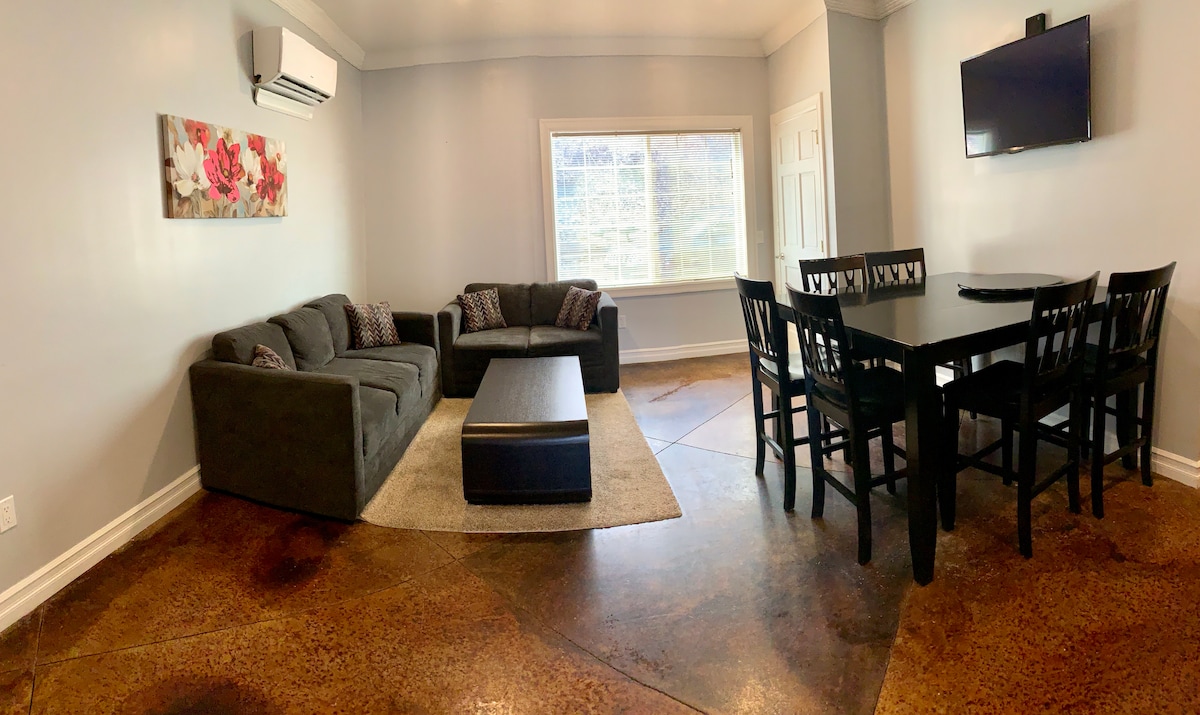 The living and dining area features a dark sectional sofa and matching coffee table set on a light rug. A dining table with six black chairs is positioned nearby. A wall-mounted TV is visible, and a large window allows natural light to fill the space.