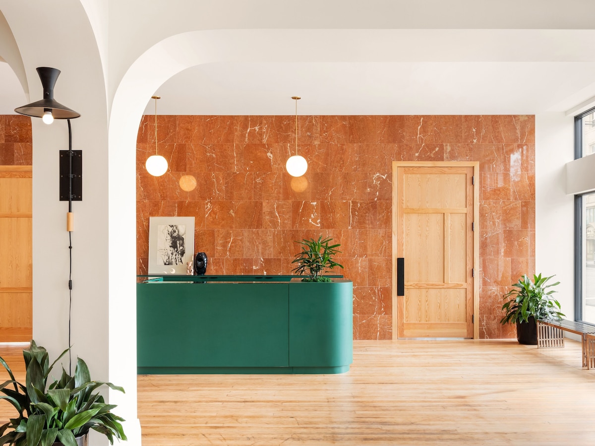 A spacious hotel lobby features a striking green reception desk against a backdrop of warm, textured orange marble walls. Soft lighting is provided by pendant lamps, while several potted plants add a touch of greenery to the inviting environment.