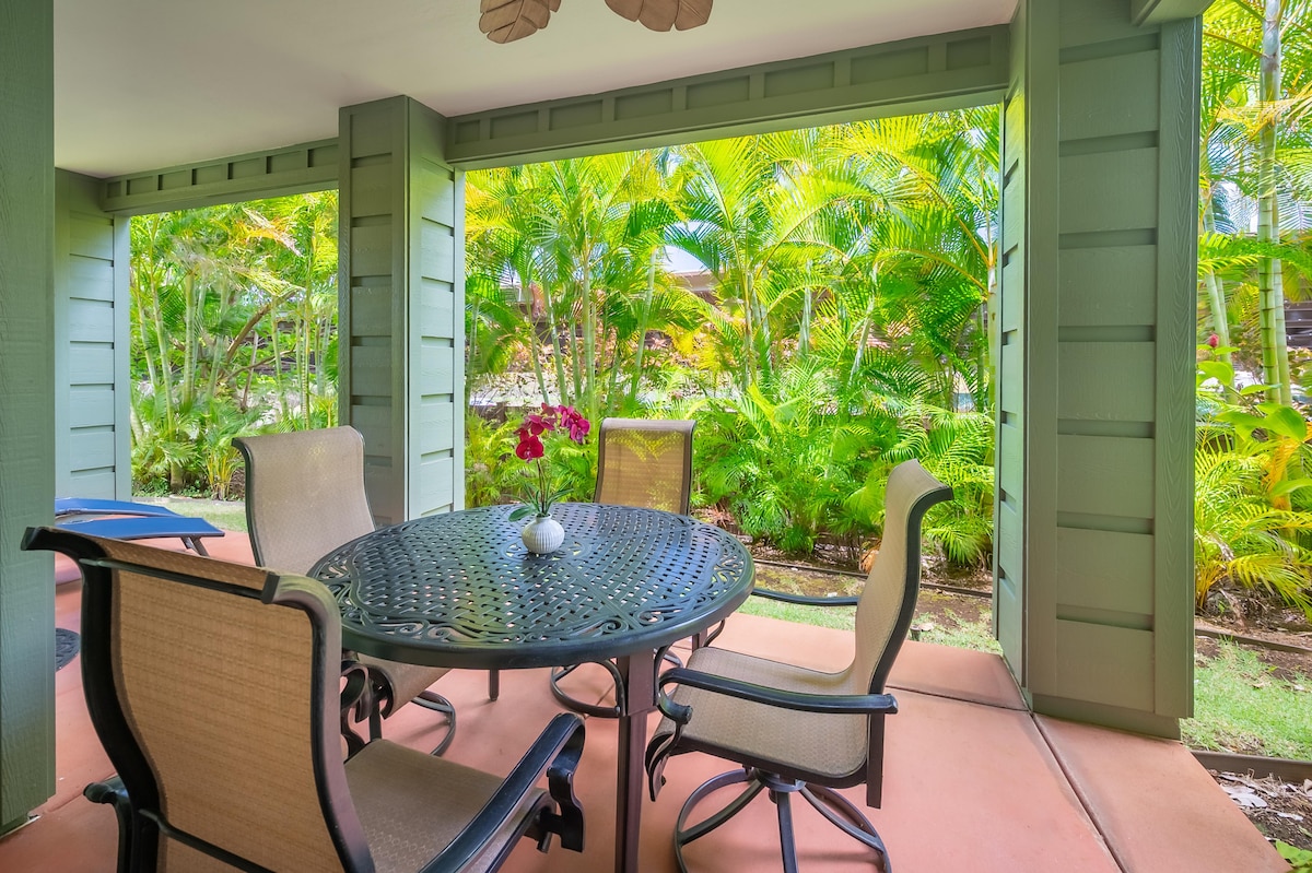 A shaded lanai features a round table surrounded by four chairs, set against a backdrop of lush tropical greenery. The vibrant plants create a serene outdoor space, complemented by a small decorative item placed center on the table.