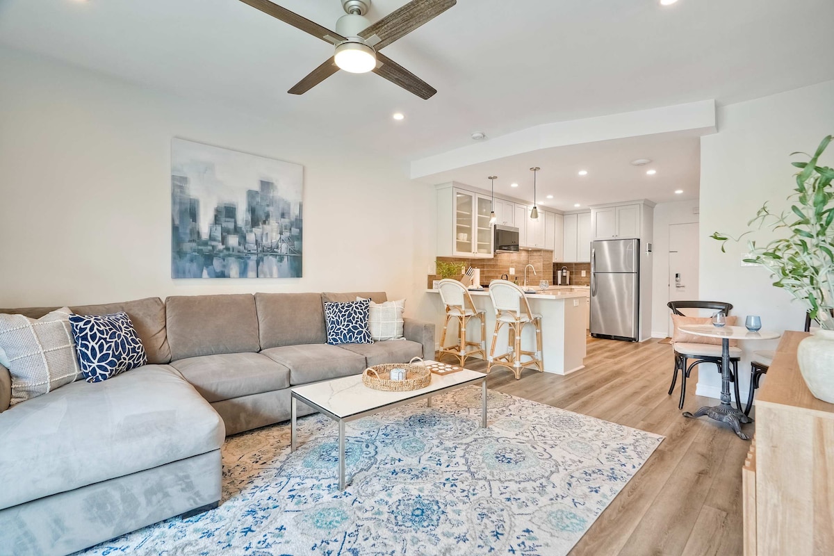 A spacious living area is highlighted by a neutral-colored sectional sofa adorned with decorative pillows. A light wood coffee table sits atop an area rug featuring a detailed pattern. The open design connects to a modern kitchen with stainless steel appliances and bar seating at two stools.