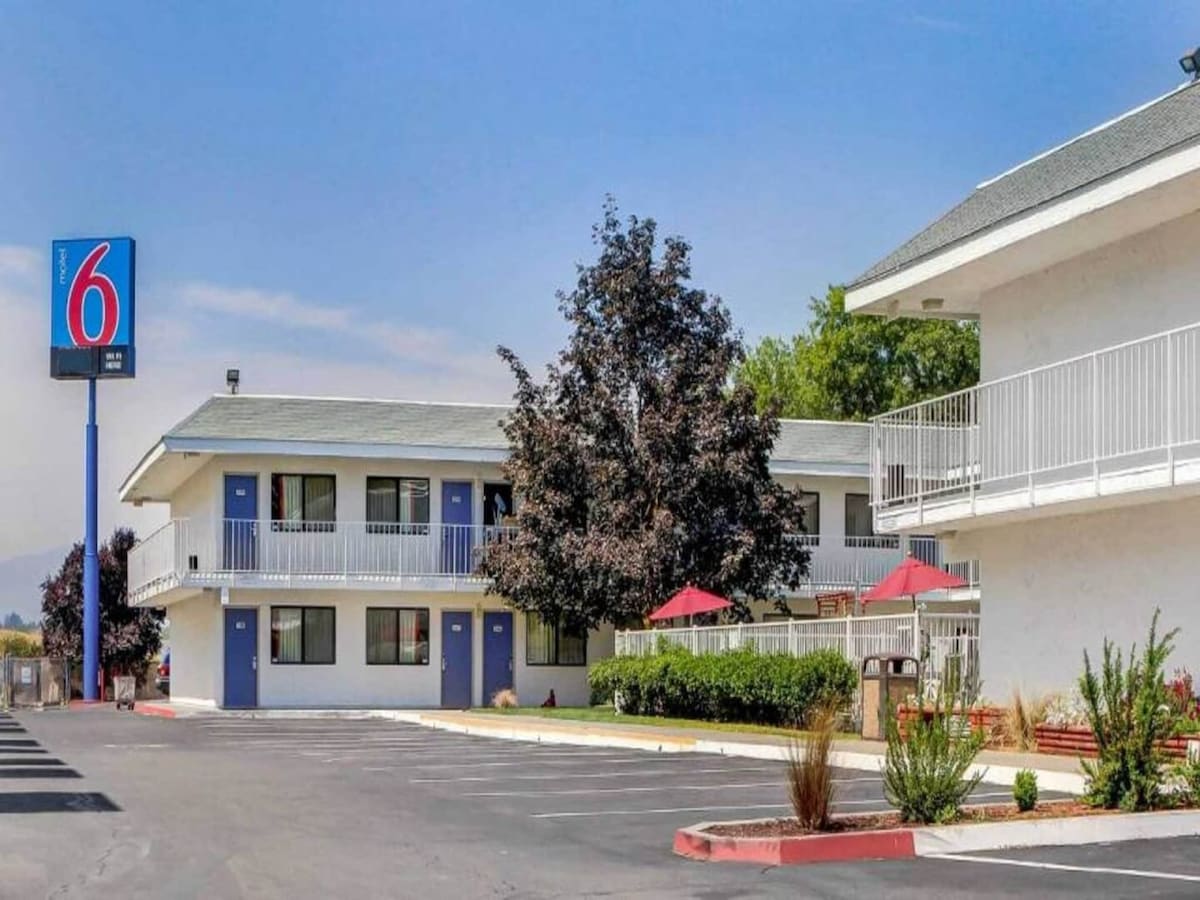 The exterior of the Motel 6 Medford displays a two-story building with blue accents and a sign featuring the number six. Surrounding shrubs and a small lawn are visible, along with outdoor seating areas under red umbrellas. Ample parking spaces are available in front.