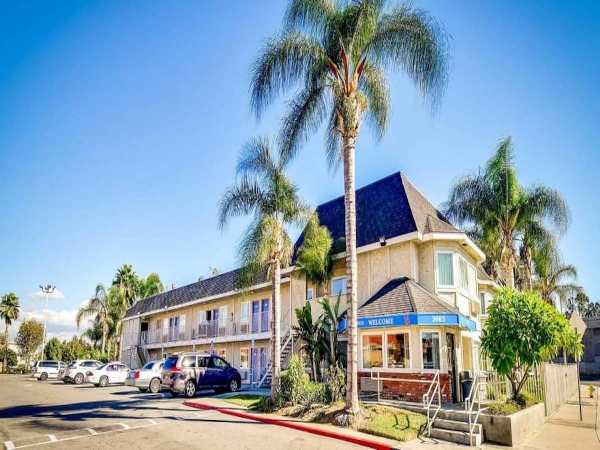 The exterior of the hotel features a two-story structure with a welcoming entrance marked by a blue sign. Lush palm trees and well-maintained landscaping surround the building. A parking lot is visible, accommodating several vehicles, and the sunny sky provides a bright backdrop.