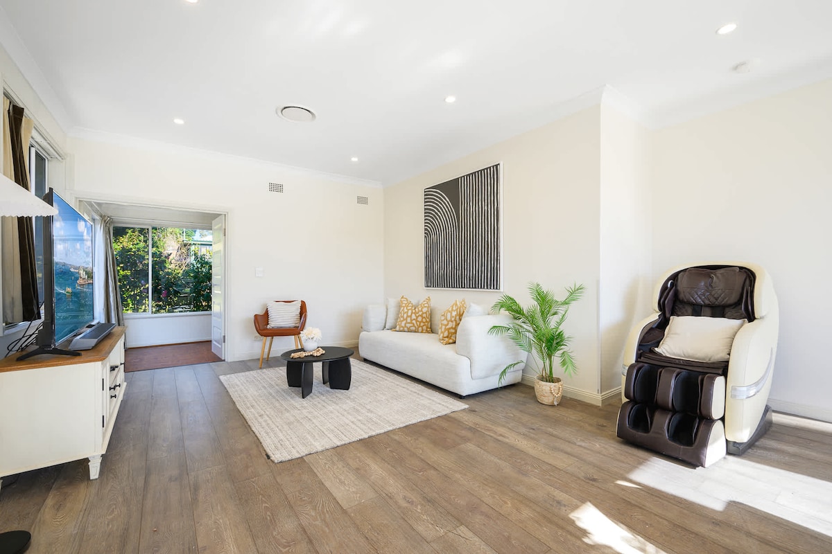 A spacious living room is presented with a comfortable sofa and a large smart TV. A massage chair is situated nearby, complemented by a small coffee table. Natural light illuminates the room, highlighting the tasteful decor and greenery from a visible outdoor space.