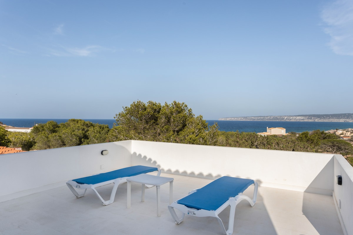 A spacious terrace offers two sun loungers with blue cushions and a small table, accompanied by a panoramic view of the Balearic Sea. The bright sky and distant coastline enhance the open and airy feel of the area.
