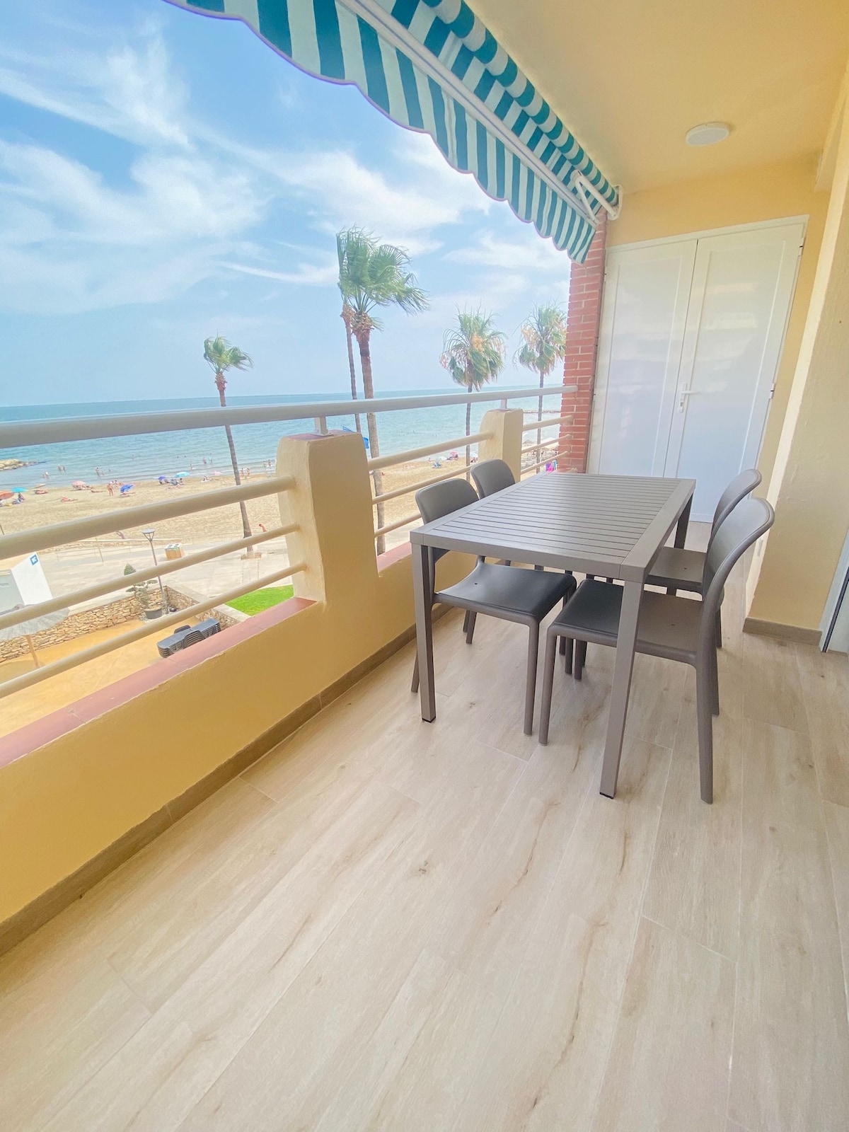 A terrace area features a table and four chairs, offering views of the beach and sea. The space is adorned with a striped awning overhead, and palm trees are visible in the distance, enhancing the coastal ambiance.