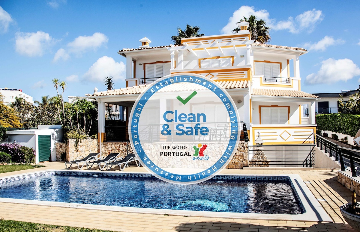 A two-story villa is presented with a well-maintained exterior and a private swimming pool in the foreground. The property features spacious balconies and a neatly landscaped garden, creating an inviting outdoor space. A 'Clean & Safe' certification seal is prominently displayed.