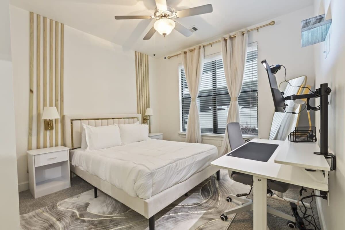 A bright bedroom features a queen-sized bed with white bedding, flanked by two nightstands with lamps. Natural light streams in through large windows dressed with light curtains. A workspace with a desk and chair is positioned nearby, complemented by a modern ceiling fan.