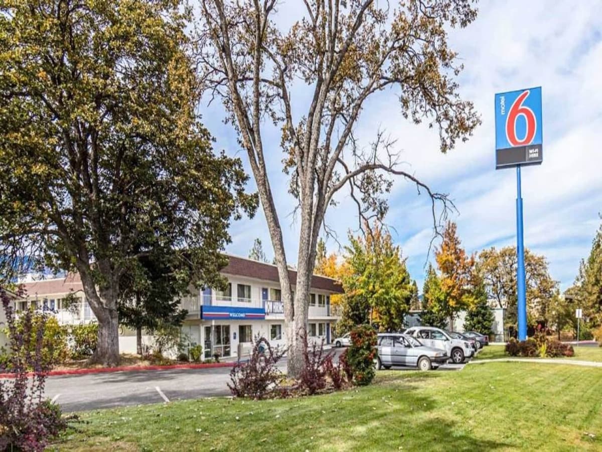 A view of the exterior of Motel 6 in Yreka, featuring a sign prominently displaying the logo. The landscape includes well-maintained grass and a variety of trees and shrubs. Vehicles are parked in the lot, indicating accessible parking options.