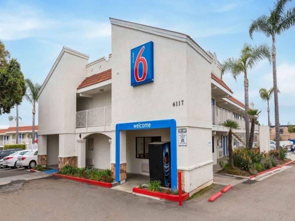 The exterior of a two-story motel features a clearly marked entrance with a large blue and red sign. Surrounding palm trees and a parking area with multiple vehicles are visible. A walkway leads to the second-floor rooms, emphasizing an inviting and accessible atmosphere.