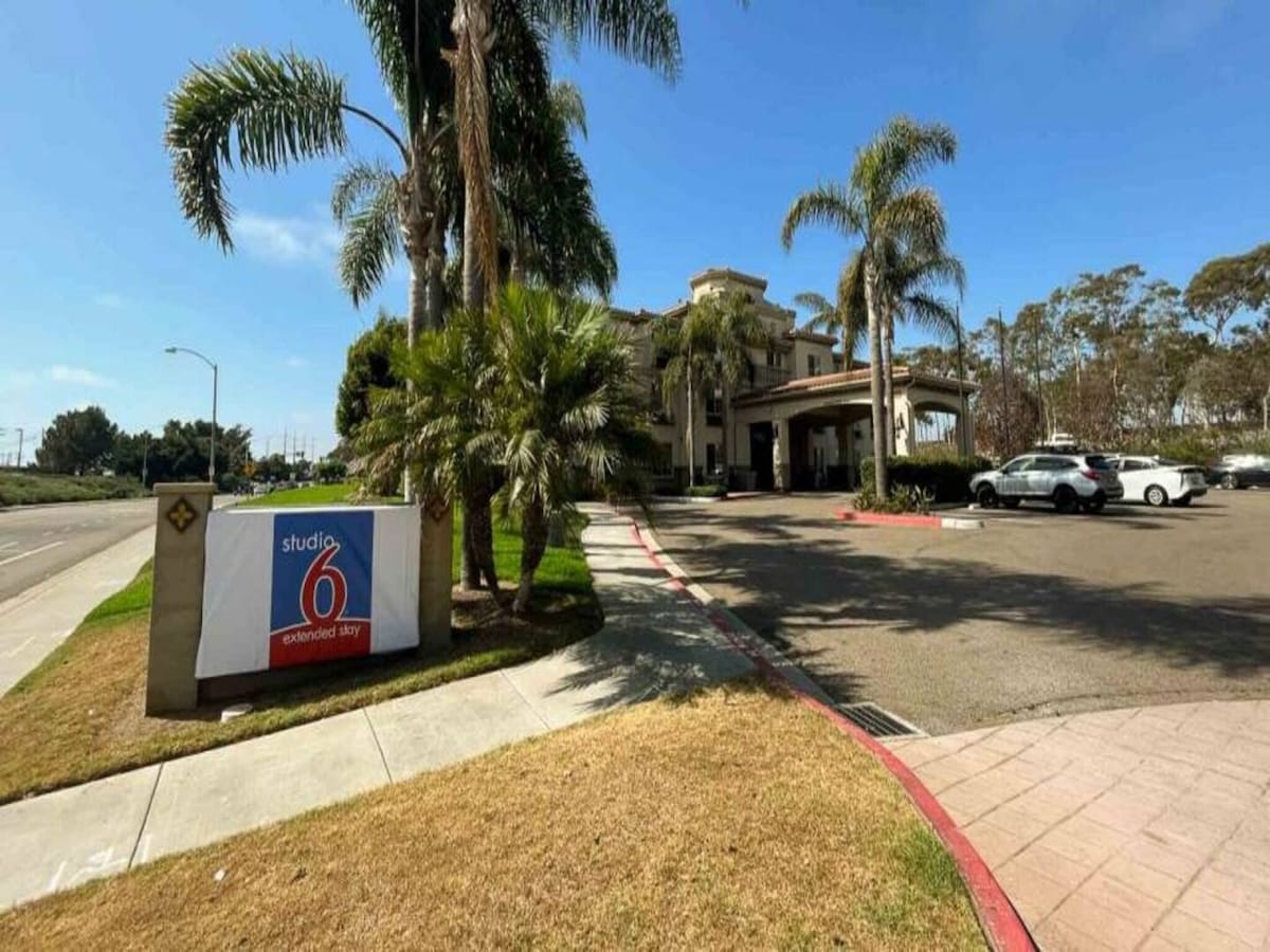 The exterior of the Studio 6 Carlsbad hotel is showcased, featuring a welcoming entrance flanked by palm trees. A large sign displaying the hotel name is visible to the left, and a parking area with vehicles can be seen in front.