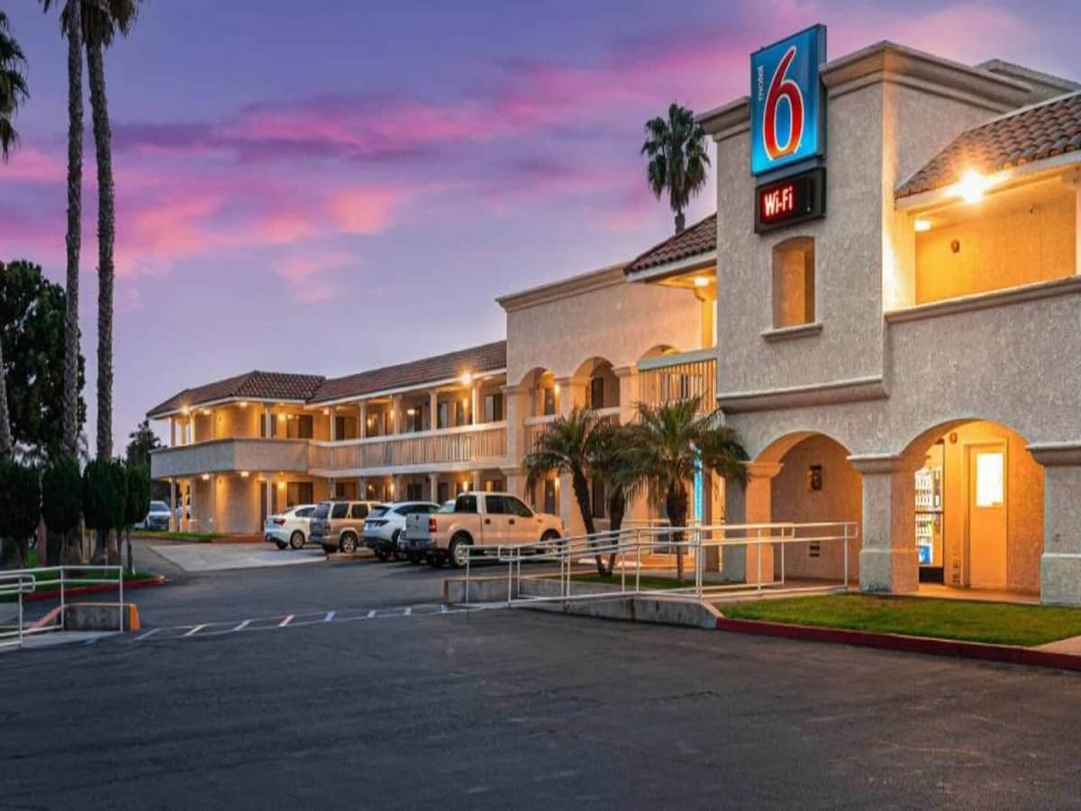 The exterior of the motel is presented, featuring a two-story building with balconies and palm trees in the foreground. Parking spaces are visible in front of the property, along with a sign indicating Wi-Fi availability. The sky is illuminated in soft hues at sunset.