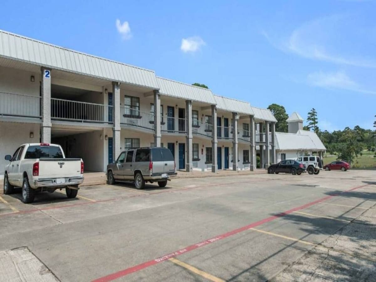 The exterior of a motel building is presented, showcasing two stories with enclosed balconies. Several vehicles are parked in a spacious lot, surrounded by greenery in the background. Clear skies enhance the welcoming appearance of the property.