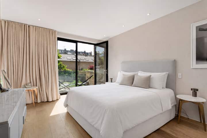 宁静的大床卧室沐浴在自然光中，配有豪华的软垫床、柔和的中性色调和落地玻璃门，可欣赏到绿树成荫的景色，营造出宁静的酒店式休憩氛围。