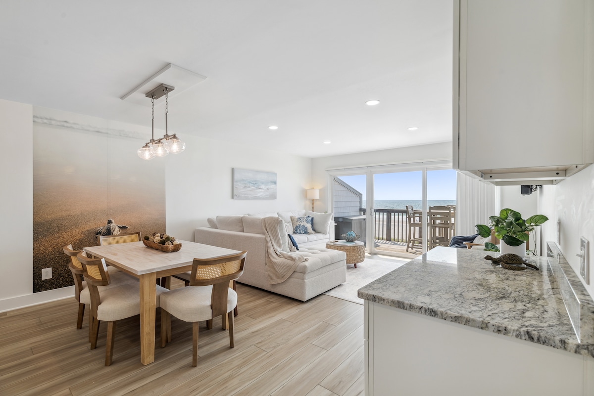 A modern living area is highlighted by a light-coloured sectional sofa, complemented by a wooden dining table with four chairs. Large sliding glass doors provide access to a balcony, allowing for natural light to fill the space and offering views of the ocean.