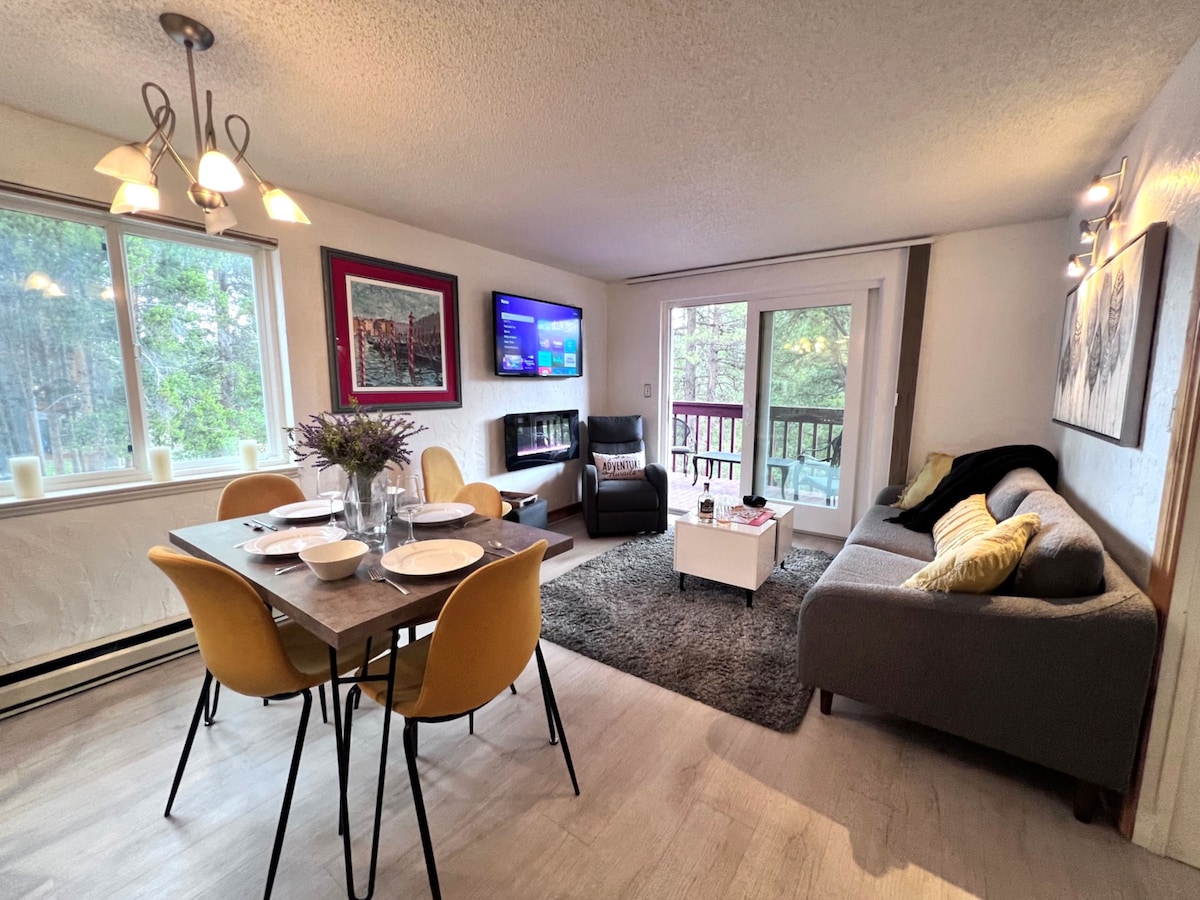 An open floor plan features a dining table set for four and a comfortable living area. A large window provides natural light and views of surrounding trees. A flat-screen TV is mounted on the wall above a modern fireplace, enhancing the cozy atmosphere.