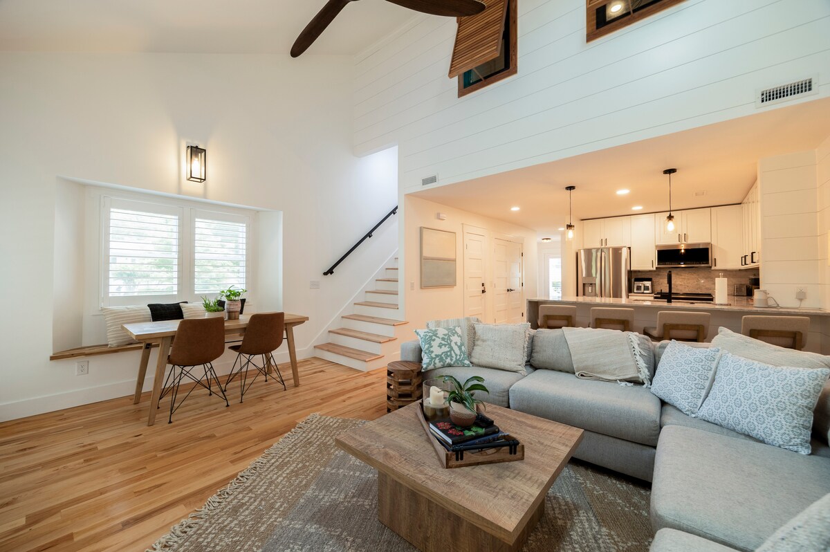 A spacious living area features a large sectional sofa paired with a wooden coffee table. Natural light fills the room, highlighting the light hardwood flooring and sleek white walls. A dining table with chairs is visible, accompanied by a staircase leading to the upper level.