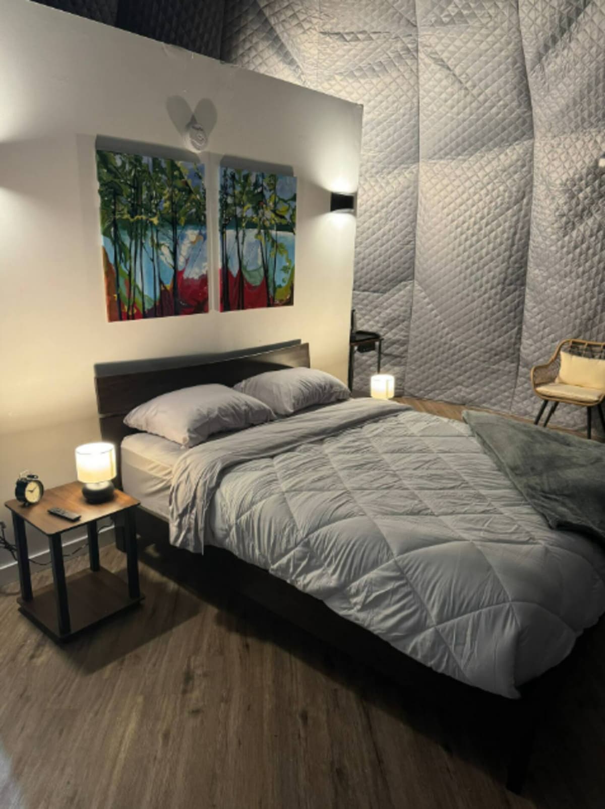 The interior of the dome features a cozy sleeping area with a queen bed dressed in soft bedding. A nightstand with a lamp and an alarm clock sits beside the bed, while colorful artwork adorns the wall. The textured walls enhance the inviting ambiance.