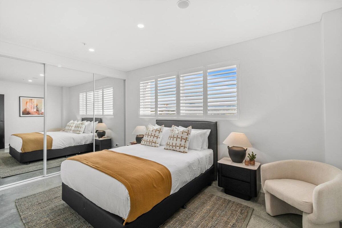 תתעוררו לאור עדין דרך חלונות סגורים בחדר השינה הראשי שלכם, הכולל מיטת קווין סייז, כורסה שיקית לקריאה שקטה וארון בגדים עם מראה לאחסון נוח.