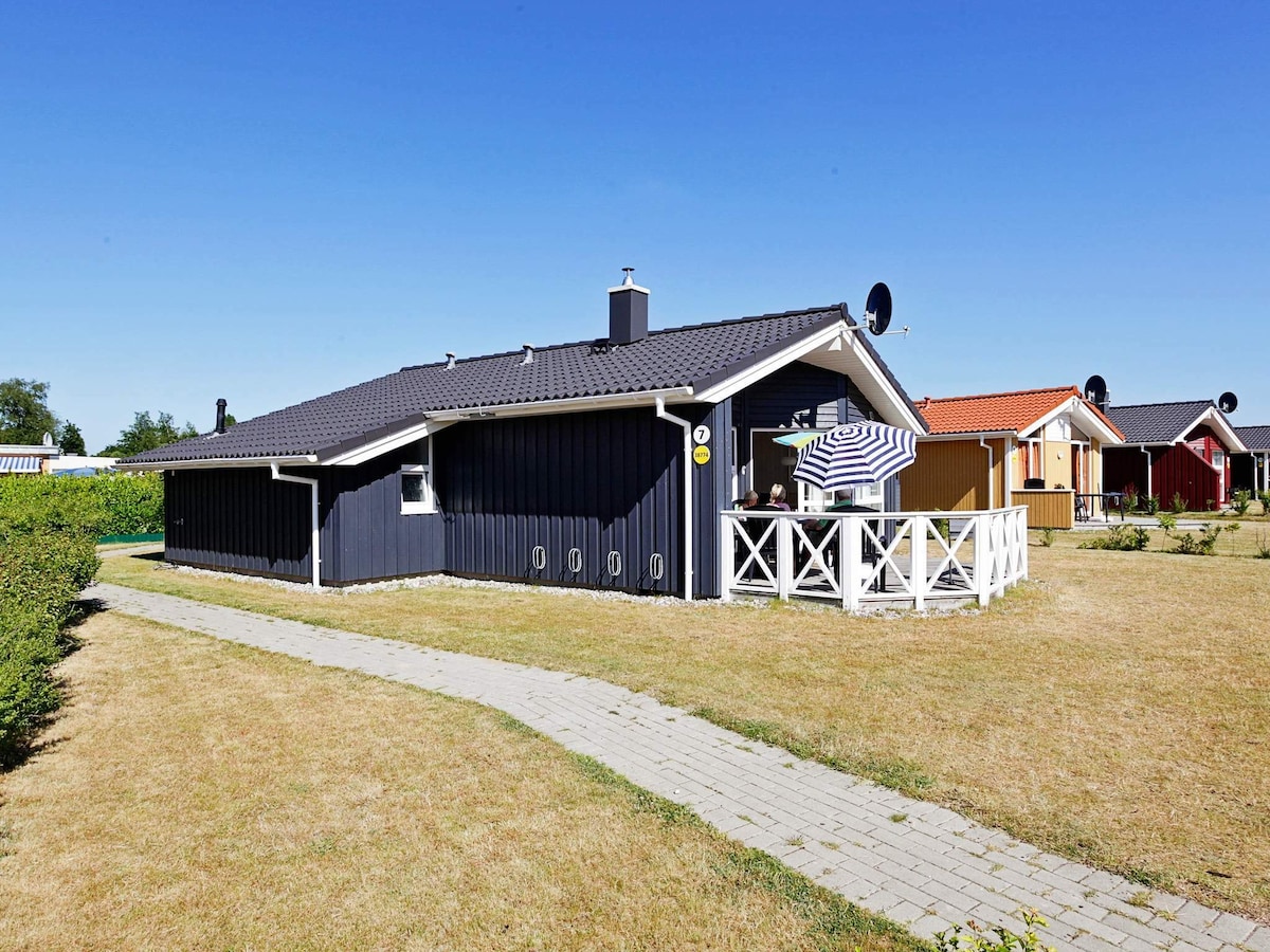 A single-story cottage features a dark exterior and a sloping roof, surrounded by a grassy area. A wooden pathway leads to the entrance, and a large terrace with a striped umbrella is visible, providing a comfortable outdoor seating area. Nearby cottages are also seen.