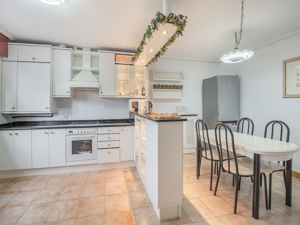 A bright kitchen area is equipped with white cabinetry, black countertops, and stainless-steel appliances. A dining table with six black chairs is positioned nearby, while natural light flows in through a window. Decorative greenery is displayed along the overhead lighting.