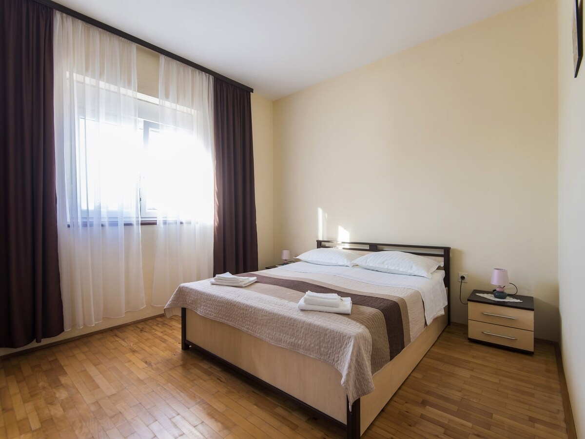 A well-lit bedroom features a spacious bed with neutral-toned bedding and two neatly arranged towels. A window covered with light curtains allows natural light to fill the room, while dark drapes provide contrast. A bedside table with a lamp is placed beside the bed.