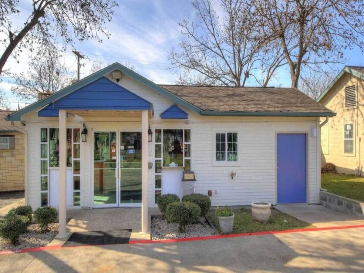 The exterior of the building is presented, featuring a blue and white facade with large windows. Landscaping includes small shrubs and planters, and a welcoming entrance is highlighted. The setting showcases a clear sky, with trees visible in the background.