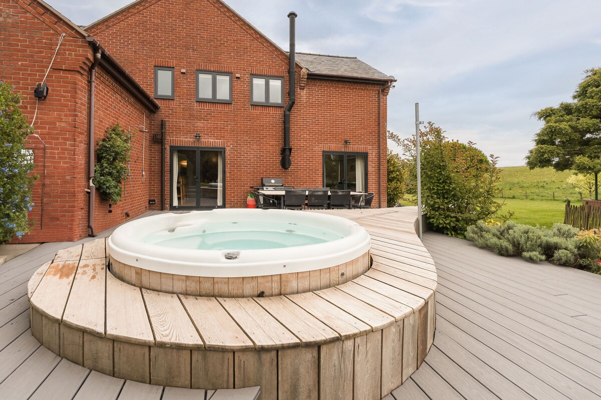A hot tub is elevated on a circular wooden platform, surrounded by a spacious deck. Two sets of double doors lead into the farmhouse, revealing outdoor dining furniture. Lush greenery and open fields are visible in the background, enhancing the peaceful ambiance.