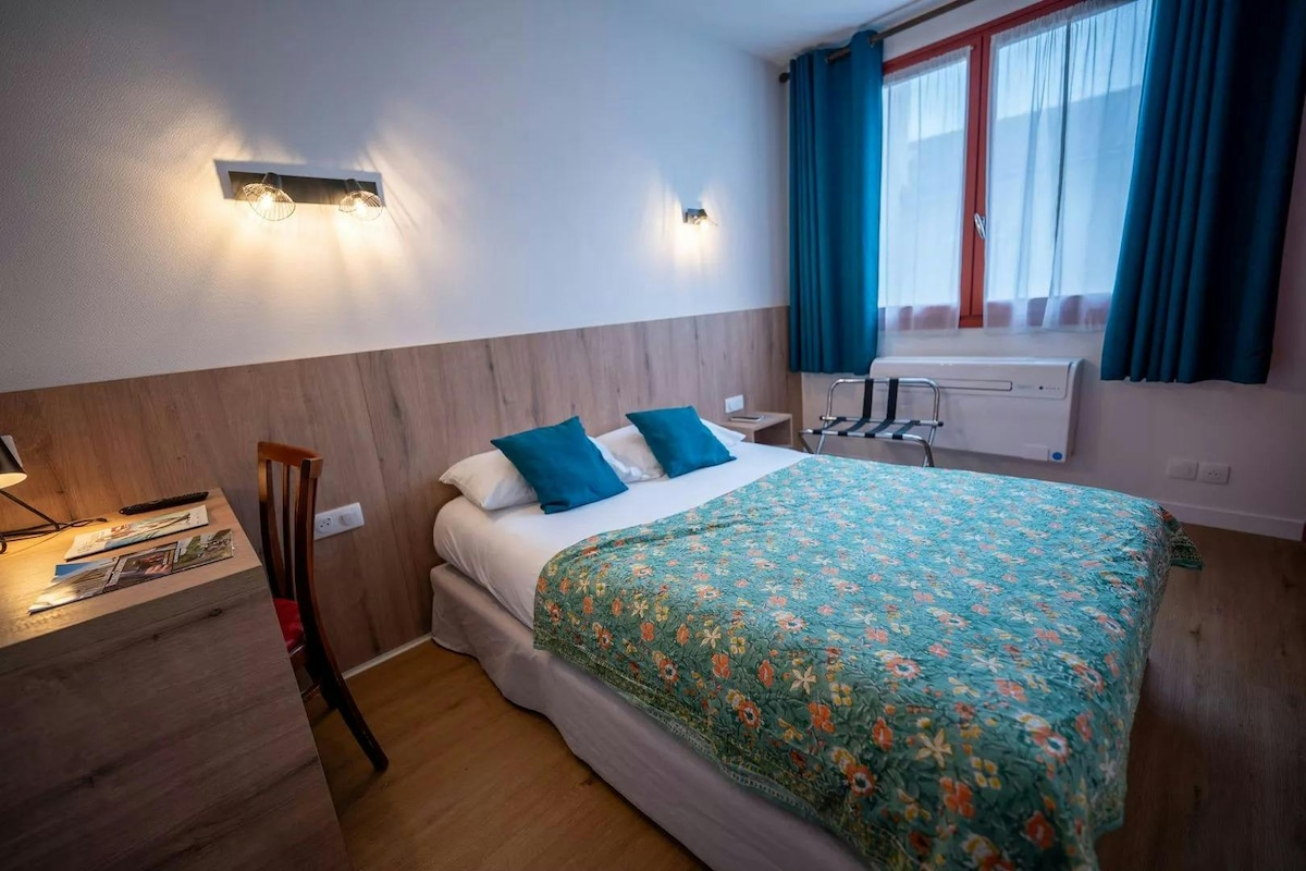 A well-appointed room features a spacious bed dressed in a floral comforter, complemented by two pillows in vibrant blue. Natural light enters through a window adorned with sheer curtains, while a wooden desk and chair are positioned nearby, providing a functional workspace.