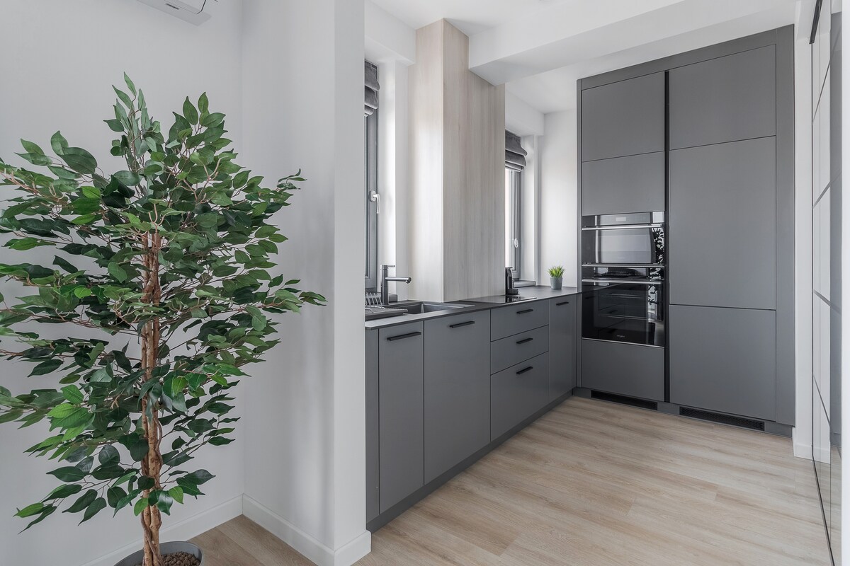 A modern kitchen features sleek gray cabinetry, with a full range of appliances including an oven and built-in microwave. Large windows provide natural light, and a small potted plant adds a touch of greenery. A clear counter space creates an uncluttered and functional area.