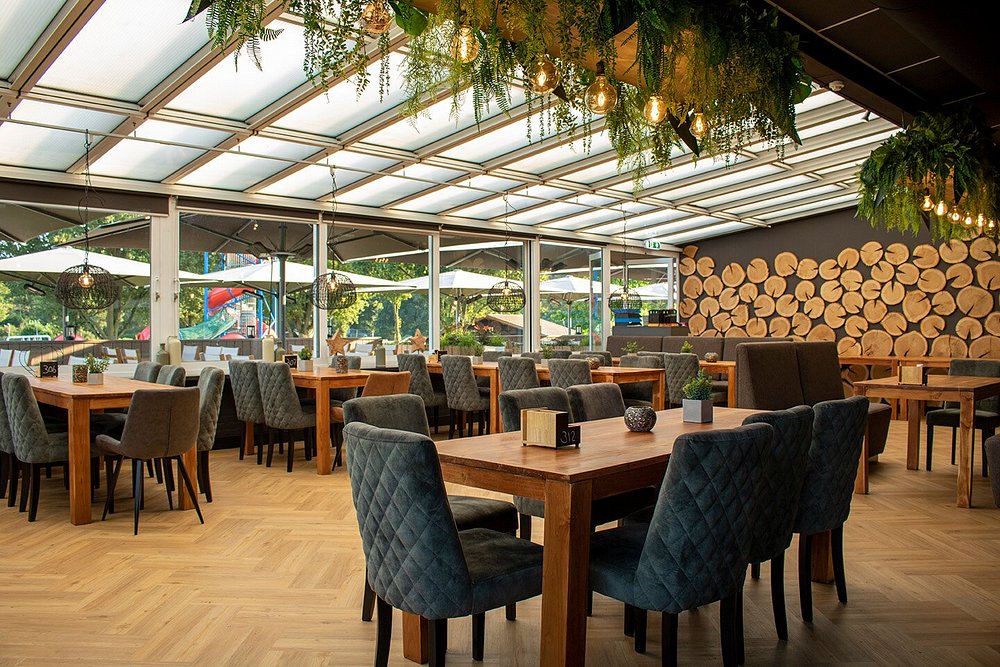 A spacious dining area is presented with wood-topped tables surrounded by upholstered chairs. Large windows allow natural light to fill the space, complemented by decorative wooden logs on the wall. Greenery hangs overhead, contributing to a relaxed atmosphere.