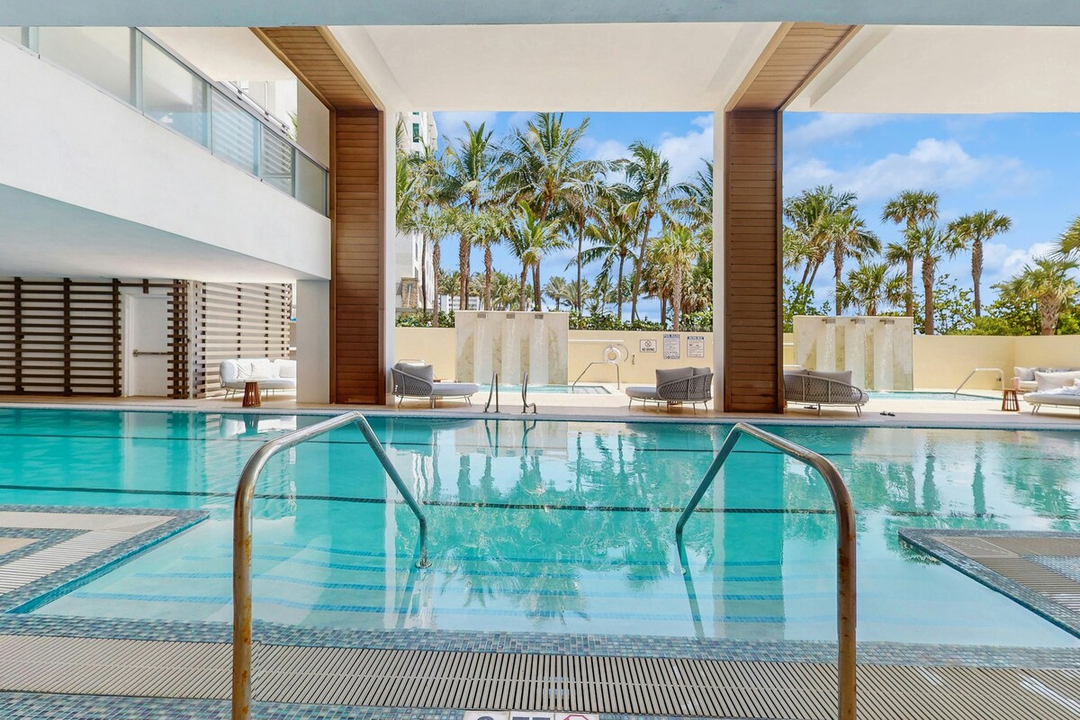 An inviting indoor pool area is featured, surrounded by a spacious deck with lounge chairs. Tall windows provide views of tropical palm trees and blue skies, creating a bright and open atmosphere. The pool’s clear water reflects the warm lighting of the space.