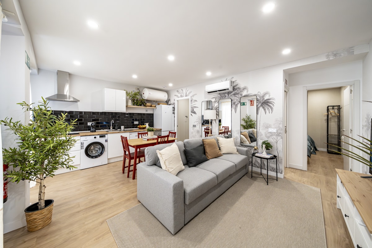 An open living area features a light grey sectional sofa positioned next to a small table and four red chairs. A kitchenette is visible with modern appliances, including a washing machine. Decorative plants are placed around the room, providing a fresh ambiance.