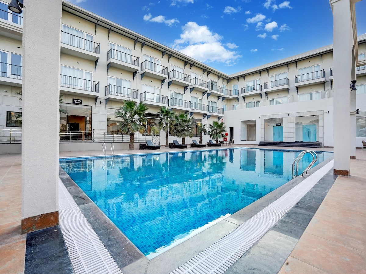 A shared swimming pool is surrounded by lounge chairs and palm trees, offering a serene area for relaxation. The hotel is designed with a modern aesthetic, featuring multiple balconies above and large windows that allow for plenty of natural light.